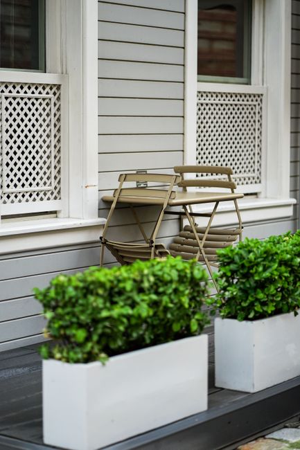 A close-up view of a garden front porch featuring a light grey wooden exterior with two double-hung windows decorated with white lace curtains behind white lattice jib windows. In the foreground, two rectangular white planters with lush, well-maintained green bushes sit on a dark grey wooden deck. Leaning against the house are several beige folding chairs stacked neatly, providing a casual seating area. The scene is well-lit with natural daylight, highlighting the textures of the wooden siding, the smooth foliage of the bushes, and the weathered surfaces of the chairs, reflecting a neat and inviting outdoor space maintained by Gardeners Camden for the Camden Market area front gardens and courtyards.